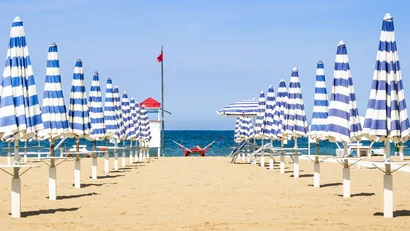 SM Hotels: ti meriti il mare tutto l’anno Ombrelloni blu e bianchi chiusi su una spiaggia sabbiosa con il mare sullo sfondo