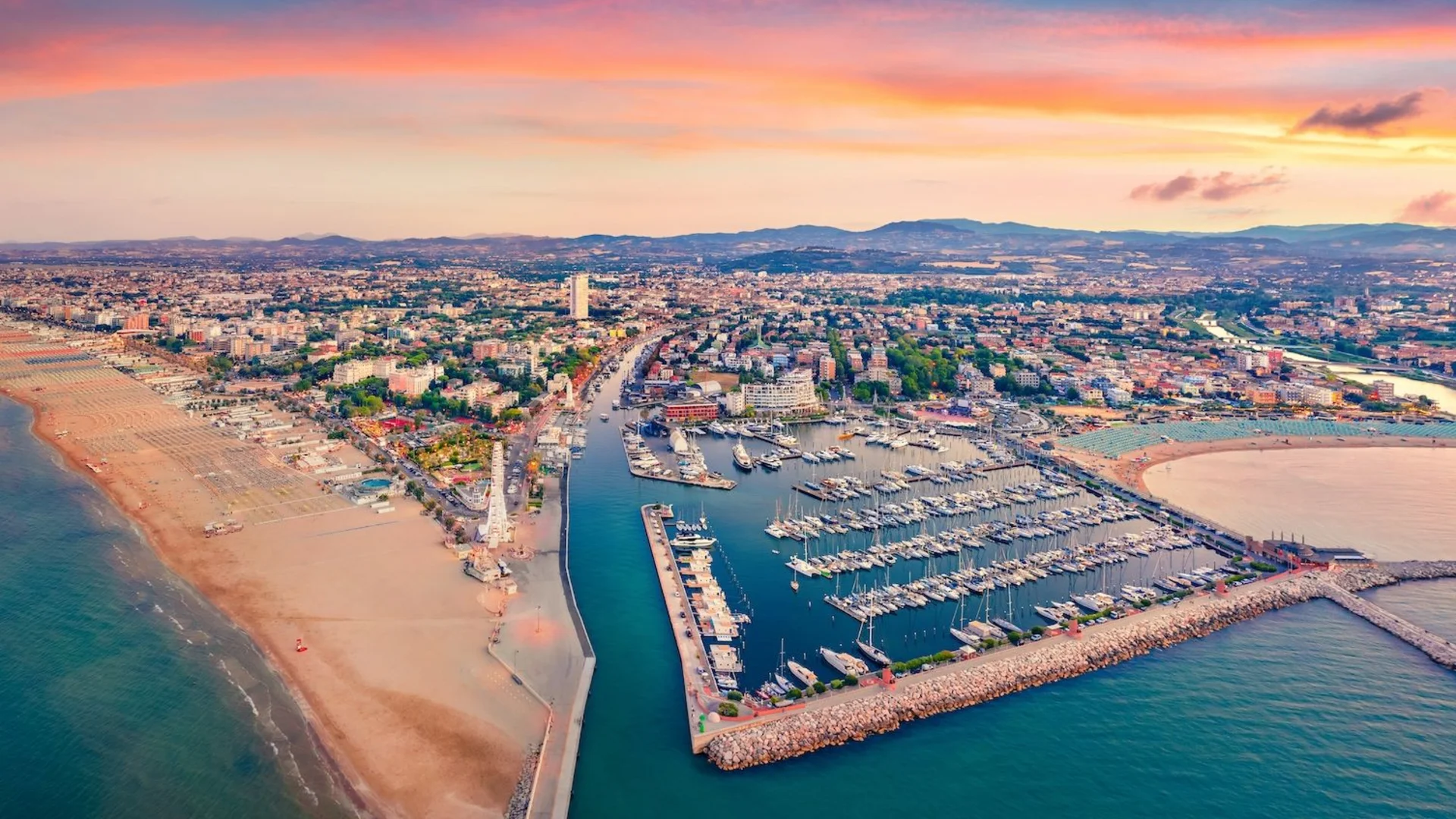SM Hotels: ti meriti il mare tutto l’anno Veduta aerea del porto e della spiaggia al tramonto con la città e le montagne sullo sfondo