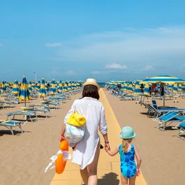 SM Hotels: ti meriti il mare tutto l’anno Donna e bambina che camminano su una spiaggia con ombrelloni blu e gialli